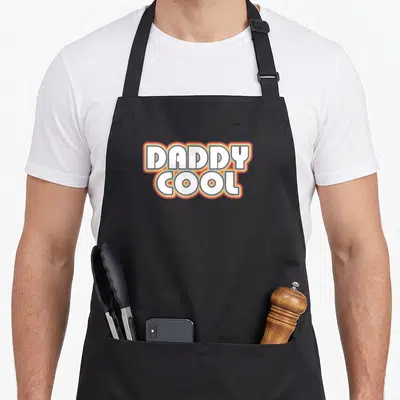 Tablier de cuisine noir avec l'inscription Daddy Cool en lettres blanches aux contours colorés style années 1970.