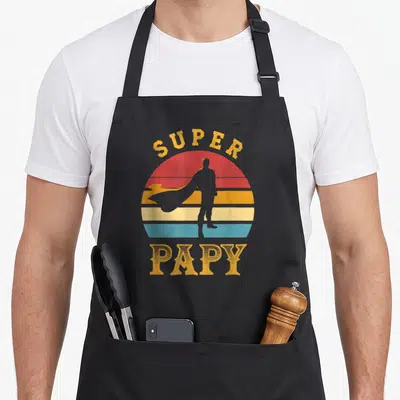 Tablier de cuisine noir avec Super Papy et silhouette de super-héros sur fond de soleil rétro multicolore.