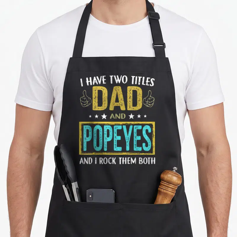 Tablier de cuisine noir avec l'inscription « I have two titles dad and Popeyes » en lettres jaunes et bleues, flanquée de deux pouces levés.