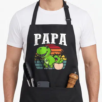 Tablier de cuisine noir avec un dinosaure vert, un bébé dinosaure et le mot PAPA en blanc sur fond de soleil coloré.