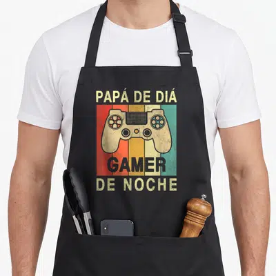 Tablier de cuisine noir avec une illustration de manette de jeu vidéo sur bandes colorées et texte en espagnol.