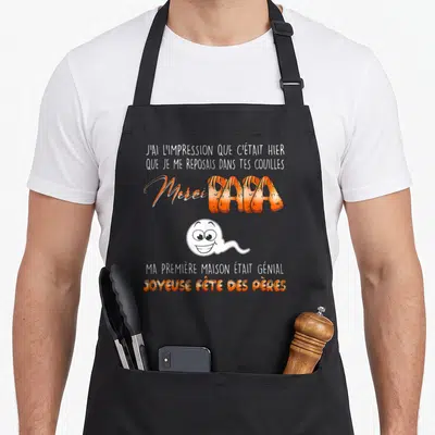 Tablier de cuisine noir avec texte orange Merci Papa et illustration de spermatozoïde souriant blanc.