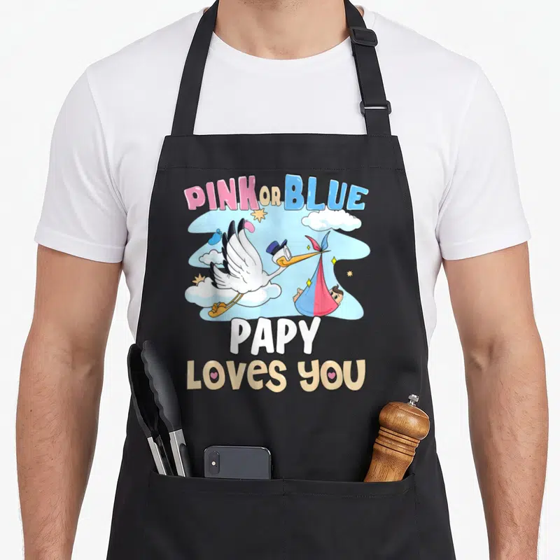 Tablier de cuisine noir avec illustration de cigogne transportant un bébé dans un drap rose ou bleu sous le texte Pink or Blue Papy Loves You.