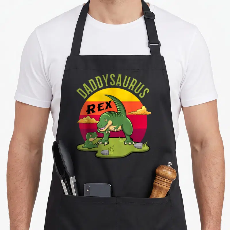 Tablier de cuisine noir avec illustration Daddysaurus Rex et dinosaure vert sur fond de soleil coloré.