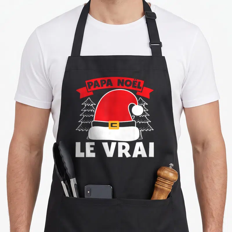 Tablier de cuisine noir avec une casquette de Papa Noël et le texte Le Vrai sur fond de sapins.