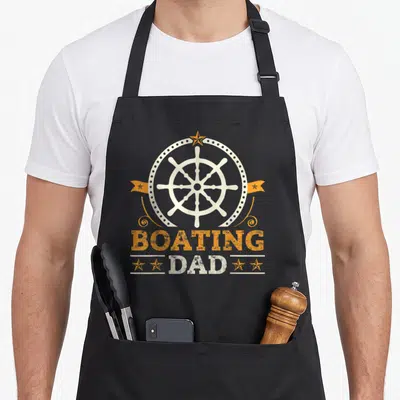 Tablier de cuisine noir avec une roue de bateau blanche, des étoiles orange et le texte Boating Dad imprimé en lettres capitales.