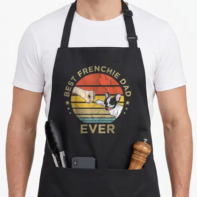 Tablier de cuisine noir avec illustration d'un bouledogue français et d'un poing humain sur fond de soleil rétro coloré.