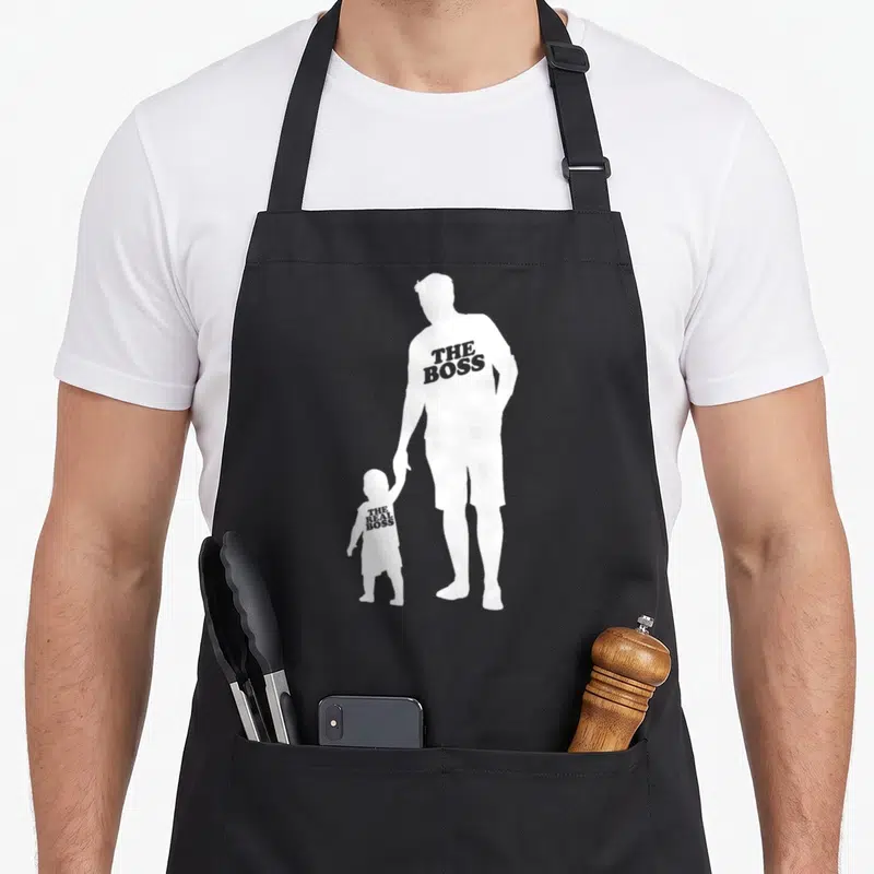 Tablier de cuisine noir avec silhouettes blanche père et enfant portant des inscriptions humoristiques sur le torse.