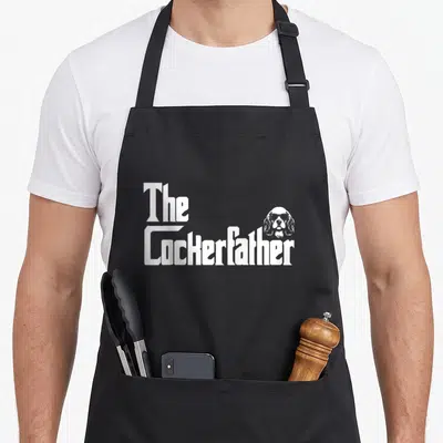 Tablier de cuisine noir avec l'inscription The Cockerfather et tête de chien aux lunettes de soleil en blanc.