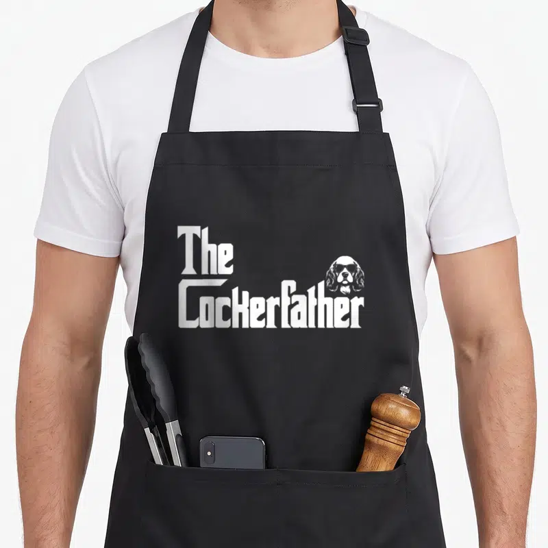 Tablier de cuisine noir avec l'inscription The Cockerfather et tête de chien aux lunettes de soleil en blanc.
