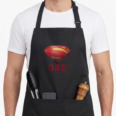 Tablier de cuisine noir avec logo Superman Man of Steel et inscription Dad en rouge.