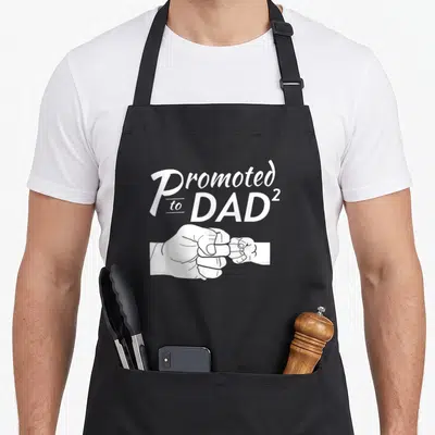 Tablier de cuisine noir avec inscription « Promoted to Dad 2 » et illustration de deux poings qui se touchent en blanc.