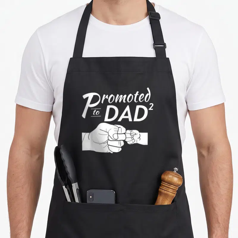 Tablier de cuisine noir avec inscription « Promoted to Dad 2 » et illustration de deux poings qui se touchent en blanc.