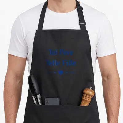 Tablier de cuisine noir avec inscription Tel Père Telle Fille et un cœur bleu encadré de flèches directionnelles.