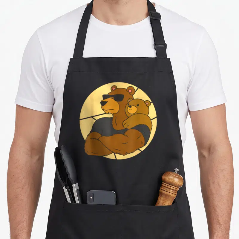 Tablier de cuisine noir avec illustration d'un ours musclé aux lunettes noires tenant un ourson dans un cercle jaune.