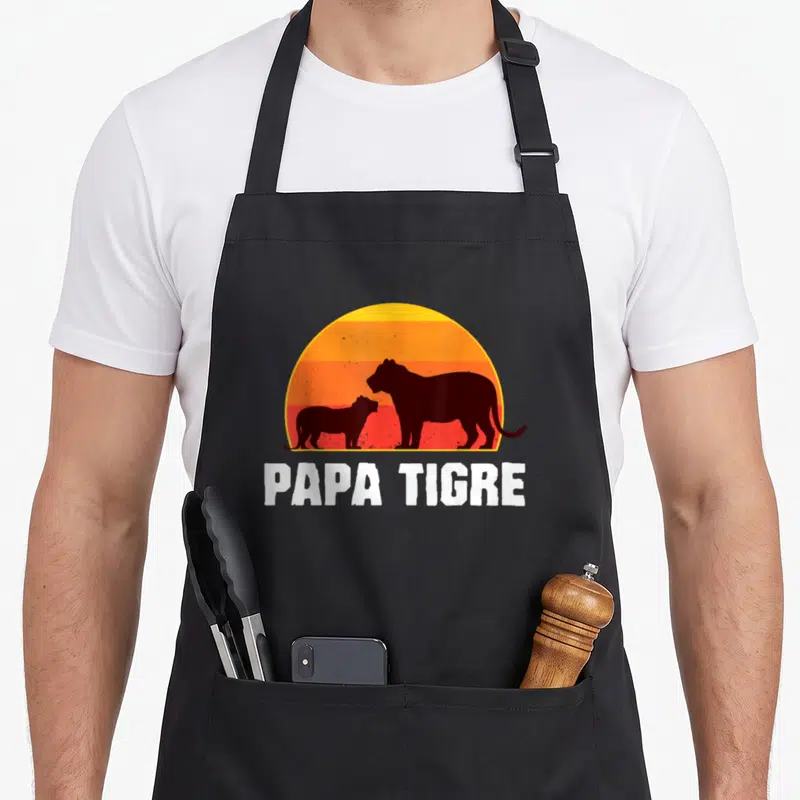 Tablier de cuisine noir avec silhouette de deux tigres et texte Papa Tigre sur fond de soleil couchant orange.
