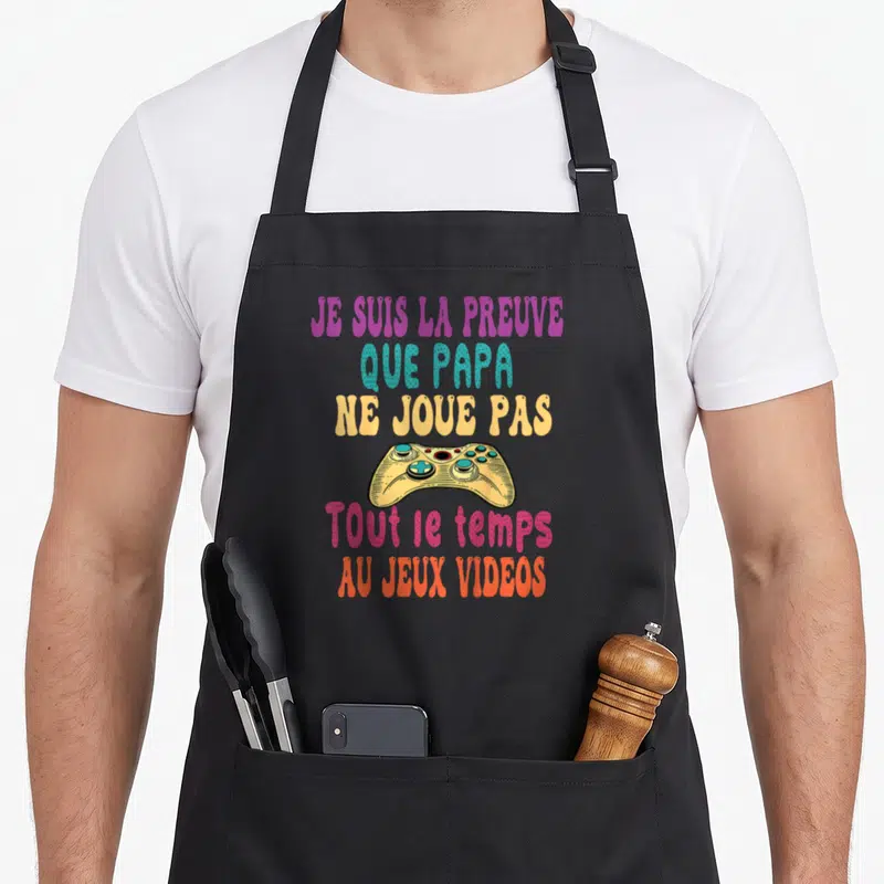 Tablier de cuisine noir avec texte multicolore et illustration de manette de jeu vidéo jaune et bleue au centre.