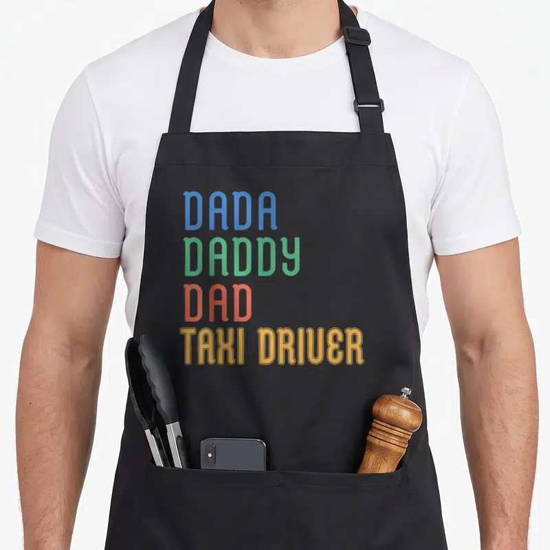 Tablier de cuisine noir avec les textes Dada Daddy Dad Taxi Driver imprimés en couleurs vives sur le devant.