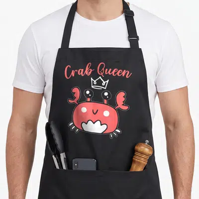 Tablier de cuisine noir avec un crabe rose souriant portant une couronne et le texte Crab Queen en haut.