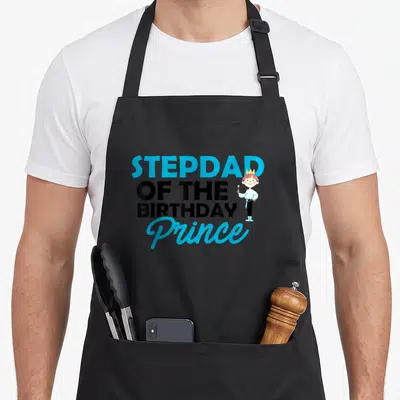 Tablier de cuisine noir avec le texte Stepdad Of The anniversaire Prince et un dessin de petit garçon portant une couronne.