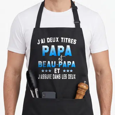 Tablier de cuisine noir avec l'inscription J'ai deux titres Papa et Beau-Papa en bleu et blanc.