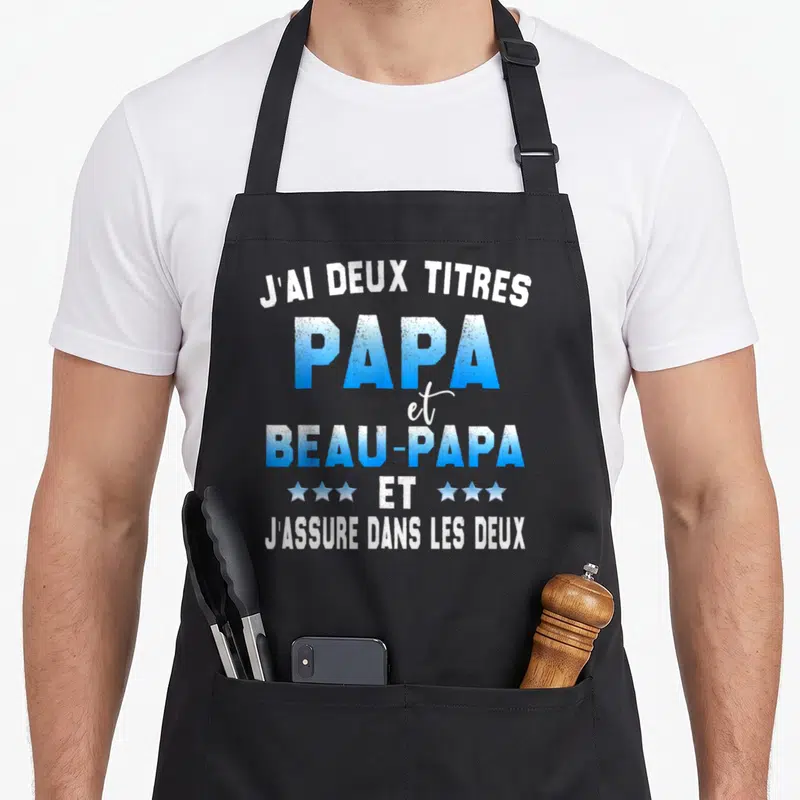 Tablier de cuisine noir avec l'inscription J'ai deux titres Papa et Beau-Papa en bleu et blanc.