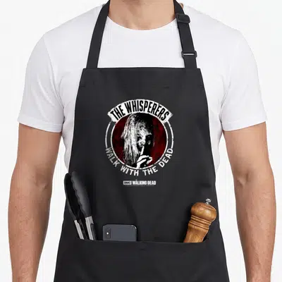 Tablier de cuisine noir avec le logo The Whisperers Walk With The Dead et un portrait stylisé au centre.
