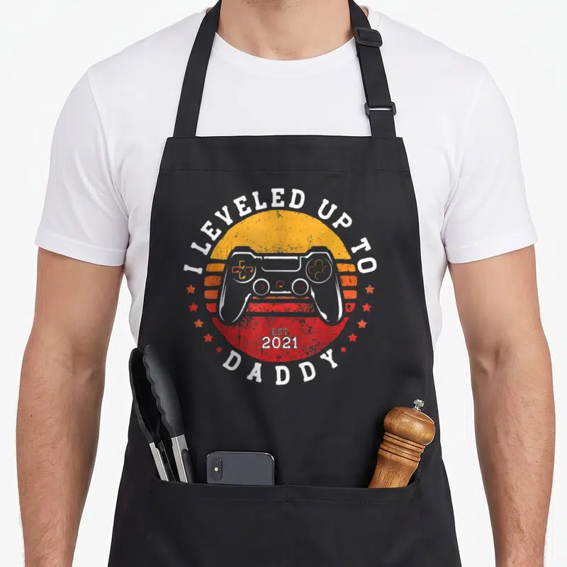 Tablier de cuisine noir avec illustration rétro de manette de jeu vidéo et texte blanc Leveled Up To Daddy 2021 Est.