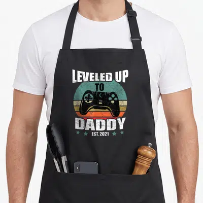 Tablier de cuisine noir avec illustration manette de jeu vidéo et texte Leveled Up To Daddy Est 2021 sur fond coloré vintage.