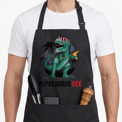 Tablier de cuisine noir avec un T-Rex vert portant des lunettes roses et tenant une perceuse jaune.