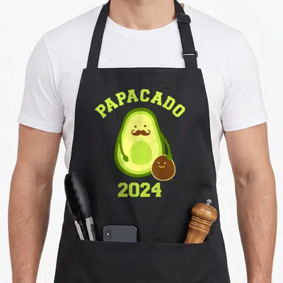 Tablier de cuisine noir avec un avocat moustachu tenant un piton souriant et le texte Papacado 2024 en vert lime.
