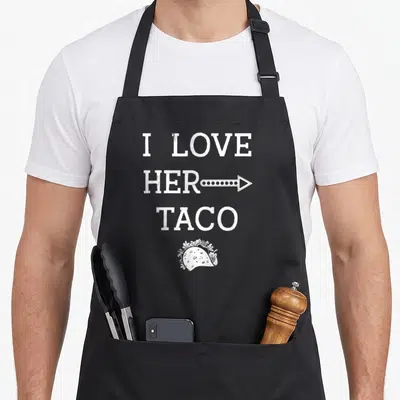 Tablier de cuisine noir avec le texte I Love Her Taco et un dessin de taco en bas du motif imprimé.
