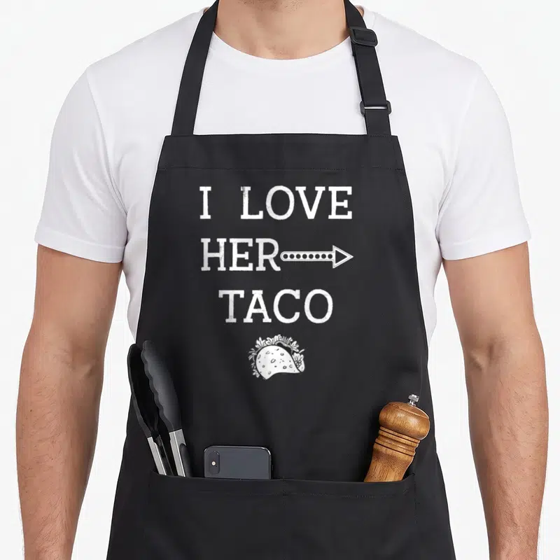 Tablier de cuisine noir avec le texte I Love Her Taco et un dessin de taco en bas du motif imprimé.