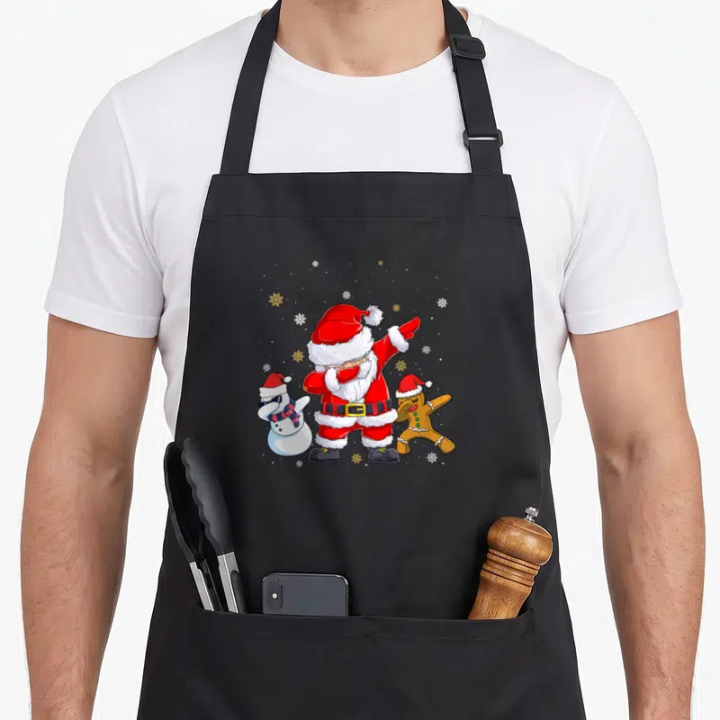 Tablier de cuisine noir avec illustration colorée d'un Père Noël faisant le dab entouré d'un snowman et d'un biscuit festif.
