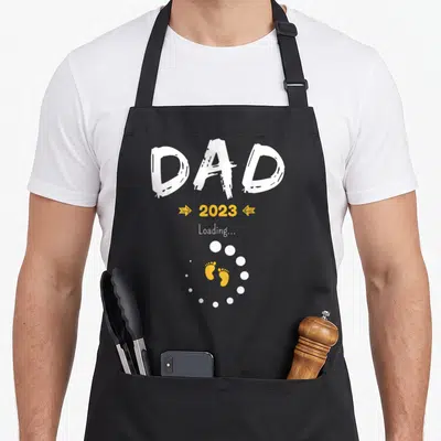 Tablier de cuisine noir avec inscription Dad 2023 Loading et empreintes de pieds jaunes au centre.