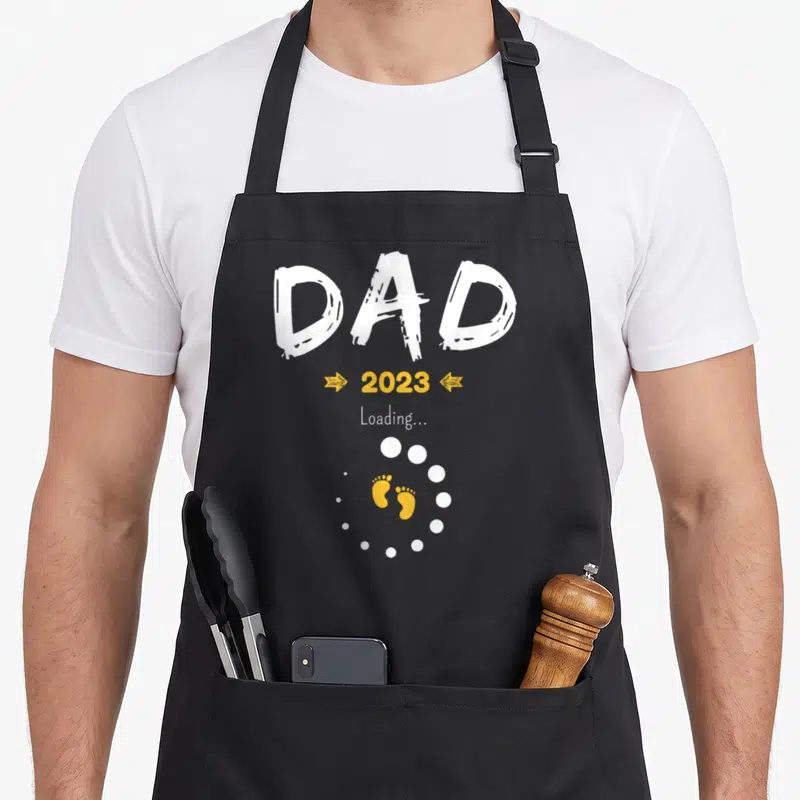 Tablier de cuisine noir avec inscription Dad 2023 Loading et empreintes de pieds jaunes au centre.