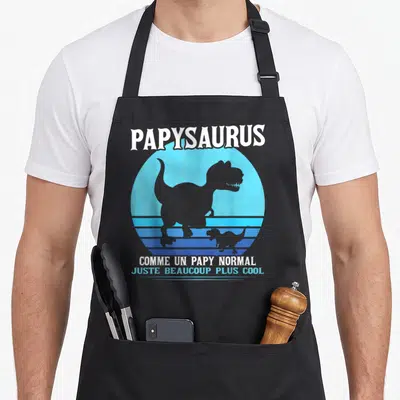 Tablier de cuisine noir avec silhouette de dinosaure T-Rex et texte blanc sur fond circulaire bleu rétro.