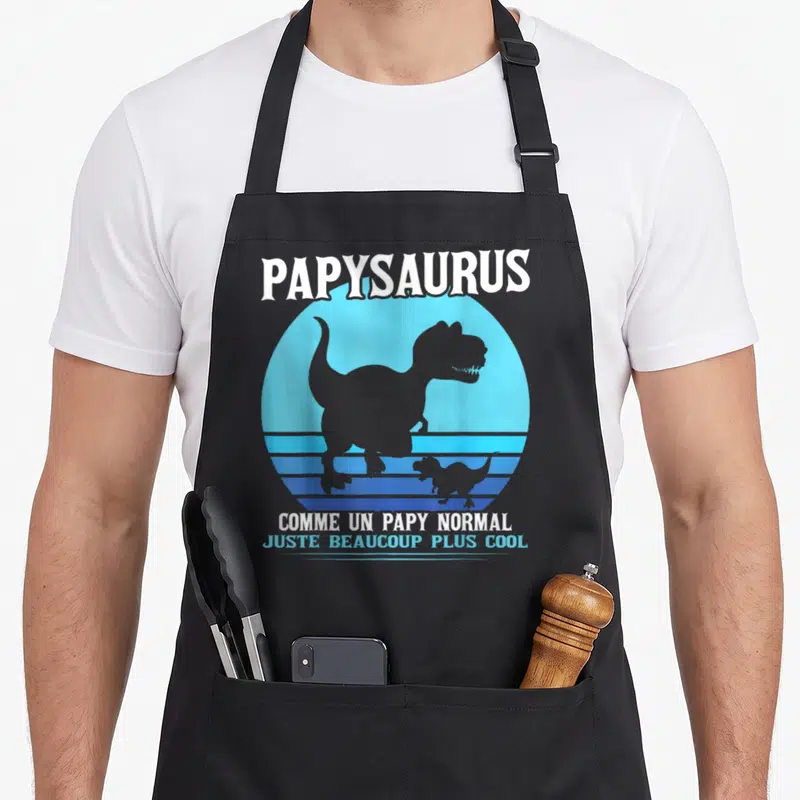 Tablier de cuisine noir avec silhouette de dinosaure T-Rex et texte blanc sur fond circulaire bleu rétro.