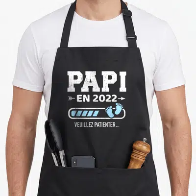Tablier de cuisine noir avec l'inscription Papi en 2022 et une barre de chargement bleue ornée de petits pieds.