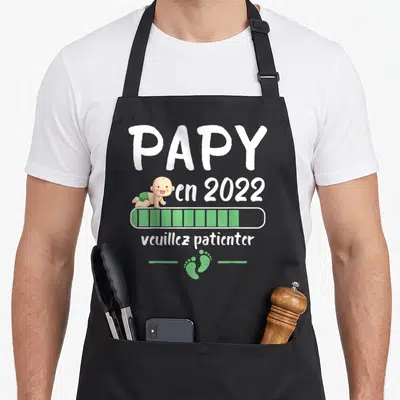 Tablier de cuisine noir avec l'inscription Papy en 2022 veuillez patienter et un dessin de bébé sur une barre de progression verte.