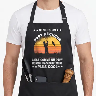 Tablier de cuisine noir avec une silhouette de pêcheur devant un soleil orange et le texte je suis un papy pêcheur plus cool en blanc.