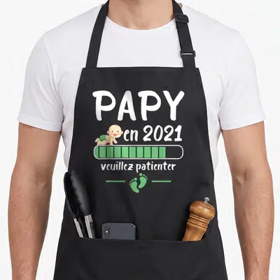 Tablier de cuisine noir avec l'inscription Papy en 2021 veuillez patienter et un bébé dessiné.
