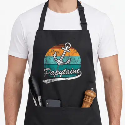 Tablier de cuisine noir avec ancre blanche vintage sur disque orange et turquoise et texte Papytaine en script blanc.