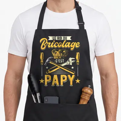 Tablier de cuisine noir avec inscription Le roi du bricolage c'est papy et outils croisés sous une couronne dorée.