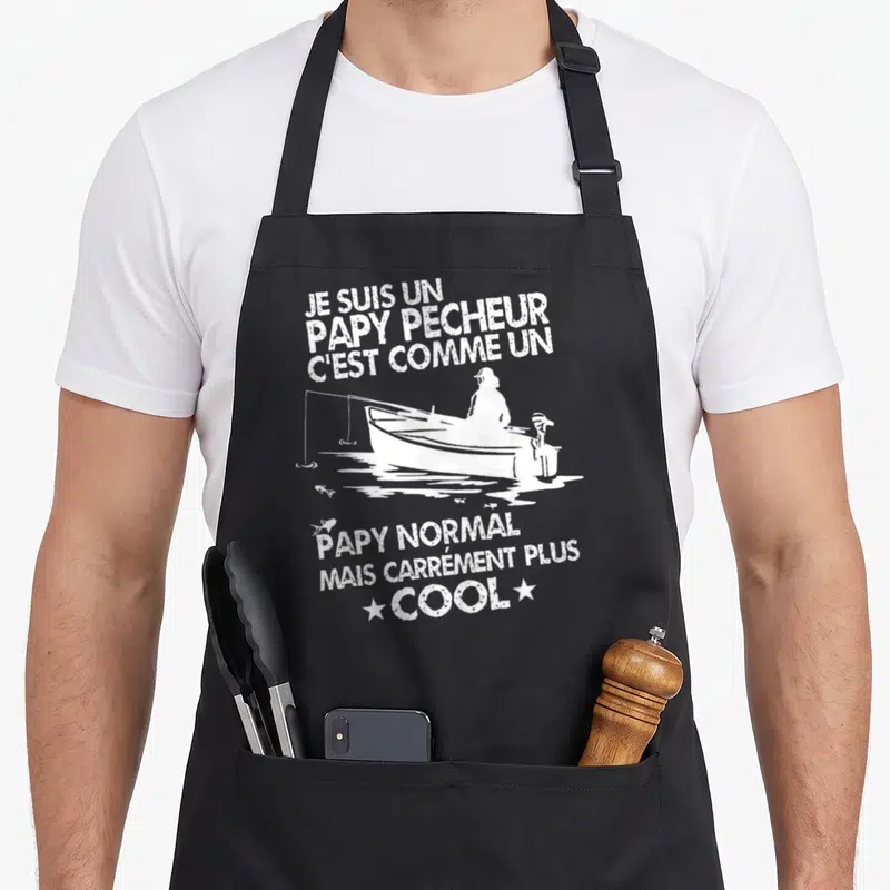 Tablier de cuisine noir avec inscription blanche sur un papy pêcheur dans une petite barque avec des poissons qui sautent.