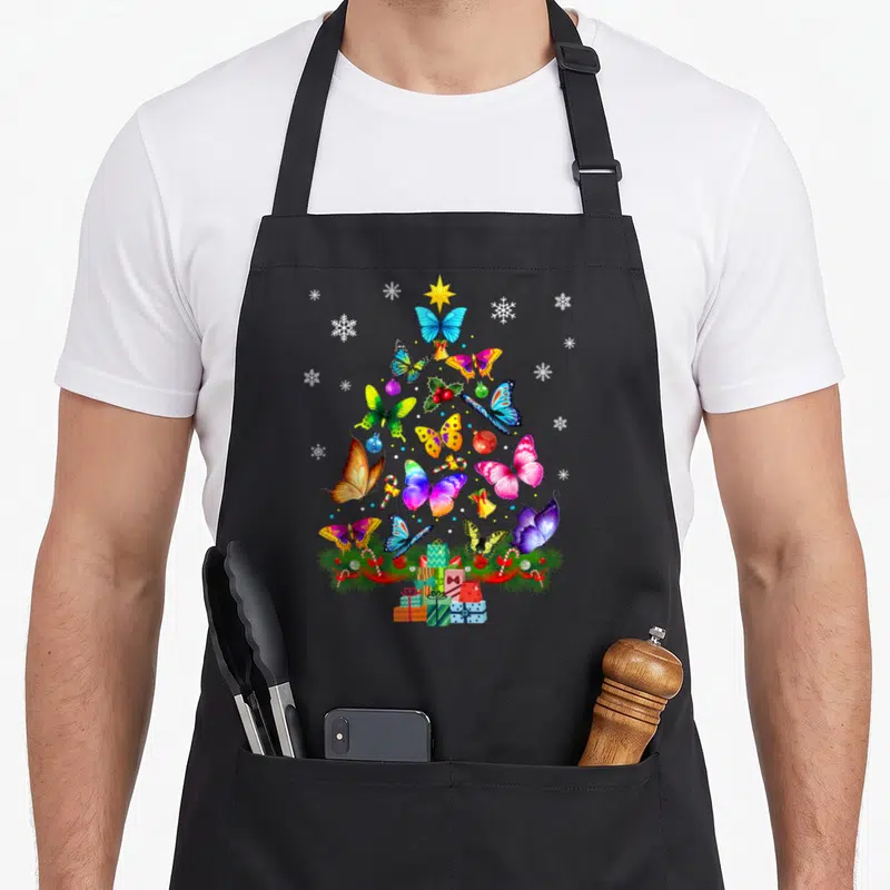 Tablier de cuisine noir avec un arbre de Noël composé de papillons multicolores, de flocons et de cadeaux à la base.