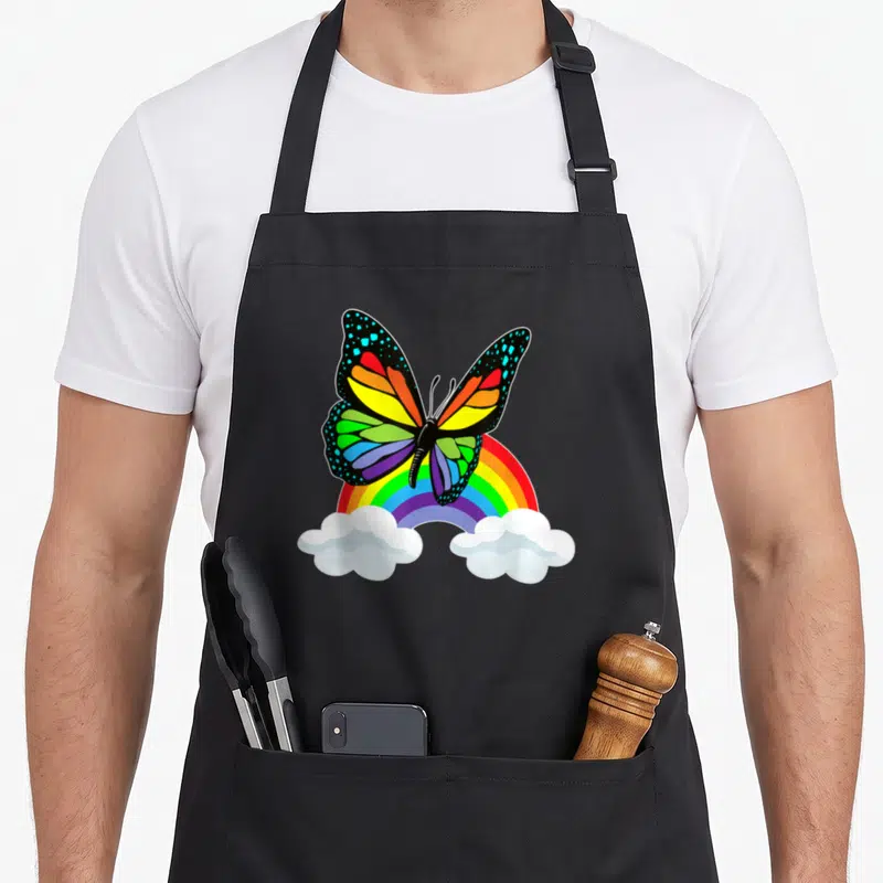 Tablier de cuisine noir avec un papillon arc-en-ciel au-dessus d'un arc de couleurs et de nuages blancs.