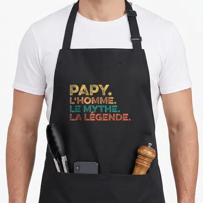 Tablier de cuisine noir avec l inscription Papy l homme le mythe la légende en lettres orange et vertes.