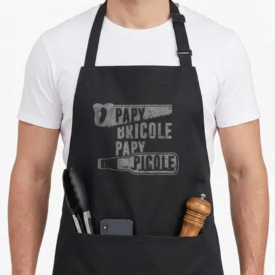 Tablier de cuisine noir avec l'inscription Papy Bricole Papy Picole et une illustration de bouteille en bas du motif imprimé.