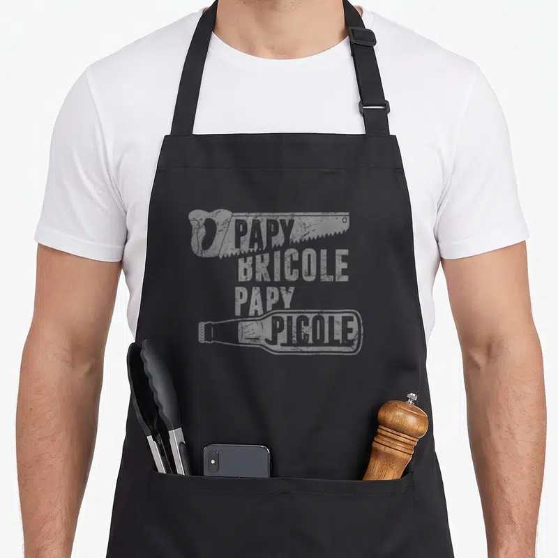 Tablier de cuisine noir avec l'inscription Papy Bricole Papy Picole et une illustration de bouteille en bas du motif imprimé.